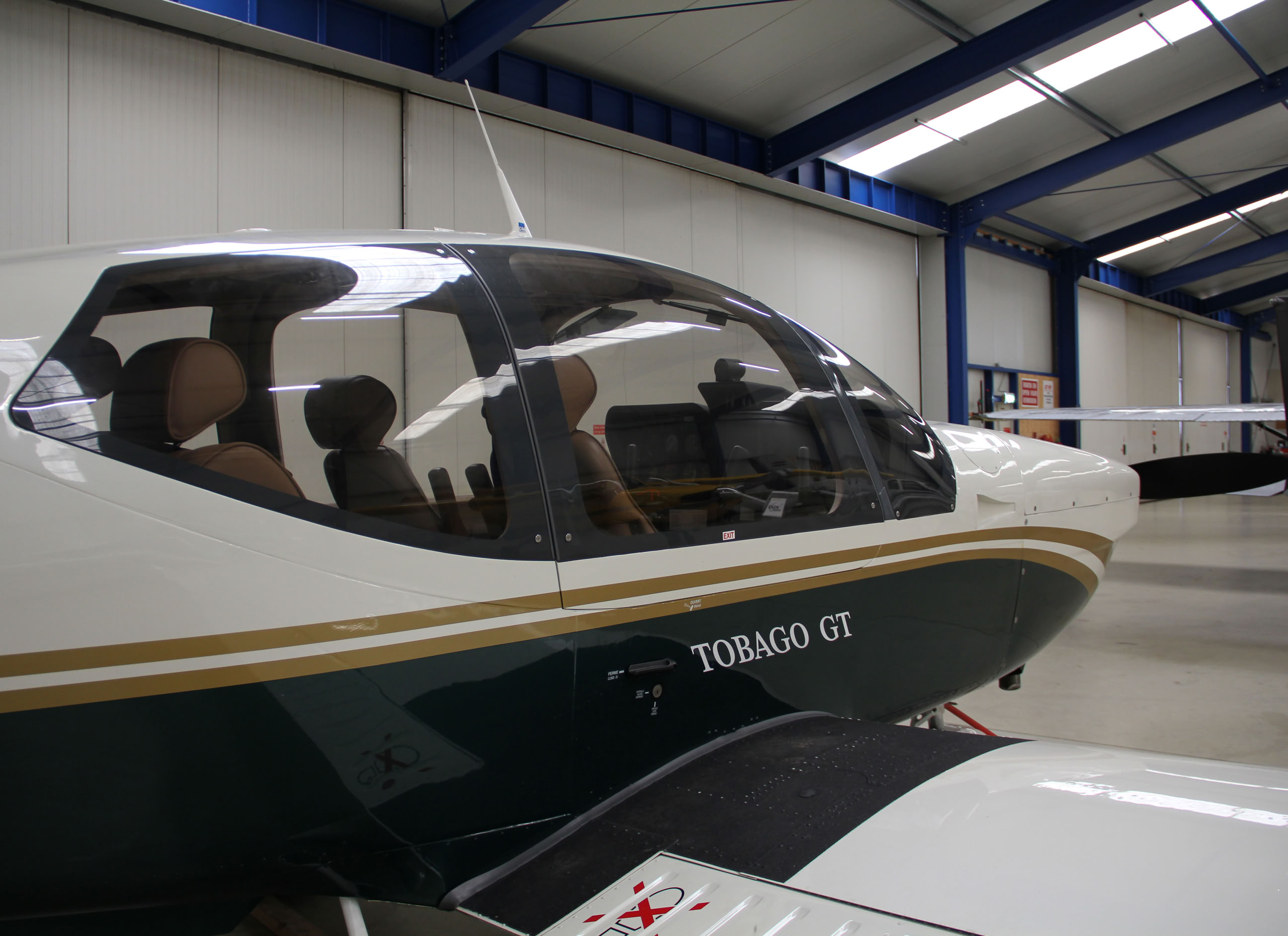 Klein vliegtuig in een hangar met blauwe stalen balken en andere vliegtuigen op de achtergrond.