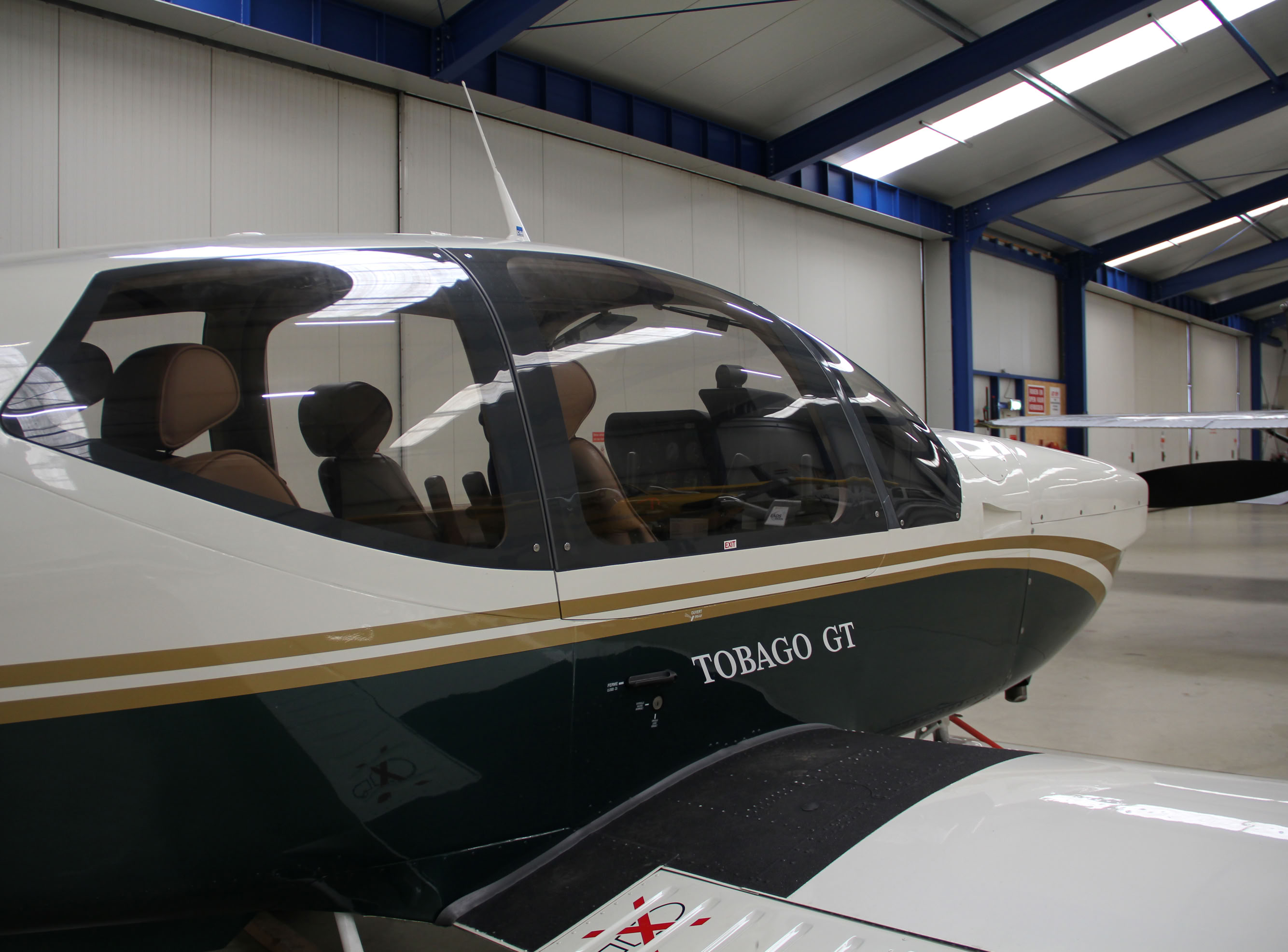 Klein vliegtuig in een hangar met blauwe stalen balken en andere vliegtuigen op de achtergrond.